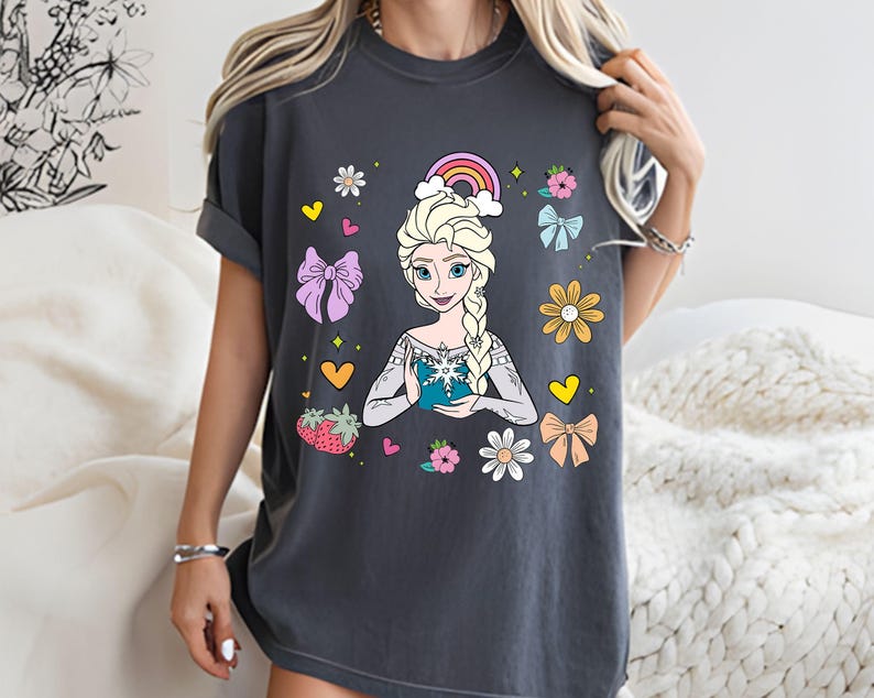 May include: A dark gray t-shirt with a colorful graphic of a cartoon princess holding a snowflake. The graphic includes a rainbow, hearts, flowers, and bows.