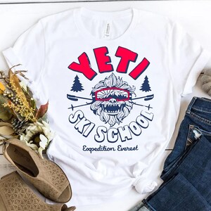 Puede incluir: Camiseta blanca con un gr&aacute;fico azul y rojo de un Yeti con gafas de esqu&iacute; y el texto "Yeti Ski School Expedition Everest".