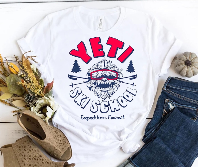Puede incluir: Camiseta blanca con un gr&aacute;fico azul y rojo de un Yeti con gafas de esqu&iacute; y el texto "Yeti Ski School Expedition Everest".