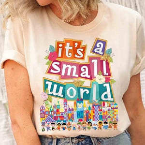 Puede incluir: Una camiseta blanca con el texto "it's a small world" en letras de colores y un gr&aacute;fico del paseo "it's a small world" en Disney World.
