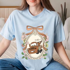 Op de afbeelding: Lichtblauw T-shirt met een cartoon afbeelding van Mater uit de film Cars. De afbeelding is omringd door een bloemmotief en een bruin lint.