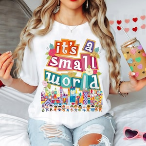 Puede incluir: Camiseta blanca con un gr&aacute;fico colorido que dice "it's a small world" y presenta una escena del paseo de Disney. El gr&aacute;fico incluye edificios coloridos, flores y personas.
