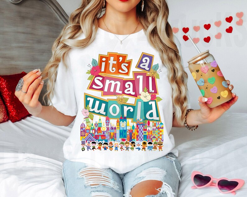 Puede incluir: Camiseta blanca con un gr&aacute;fico colorido que dice "it's a small world" y presenta una escena del paseo de Disney. El gr&aacute;fico incluye edificios coloridos, flores y personas.