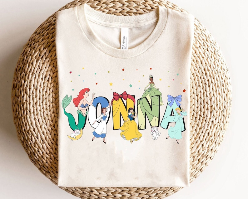 May include: A white t-shirt featuring a colourful design with Disney princesses and the name "JONNA". The princesses are dressed in their signature colours and are surrounded by stars and glitter.