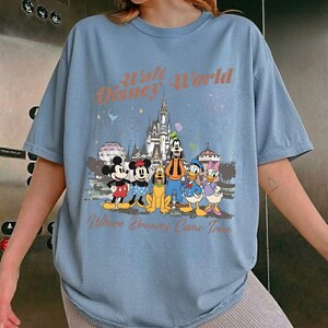 May include: Light blue t-shirt featuring a graphic of Disney characters including Mickey Mouse, Minnie Mouse, Goofy, Pluto, Donald Duck, and Daisy Duck. The shirt has the text "Walt Disney World" and "Where Dreams Come True".