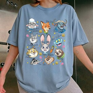 May include: Light blue t-shirt featuring a colorful graphic of various cartoon animal characters. The design includes a sheep, fox, buffalo, rabbit, and sloth, among others. The shirt is a relaxed fit.