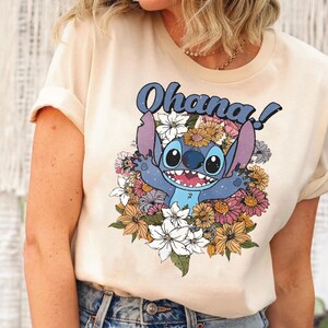 May include: A light beige t-shirt with a blue Stitch character from Lilo & Stitch surrounded by colorful flowers. The text "Ohana!" is printed in black above the character.