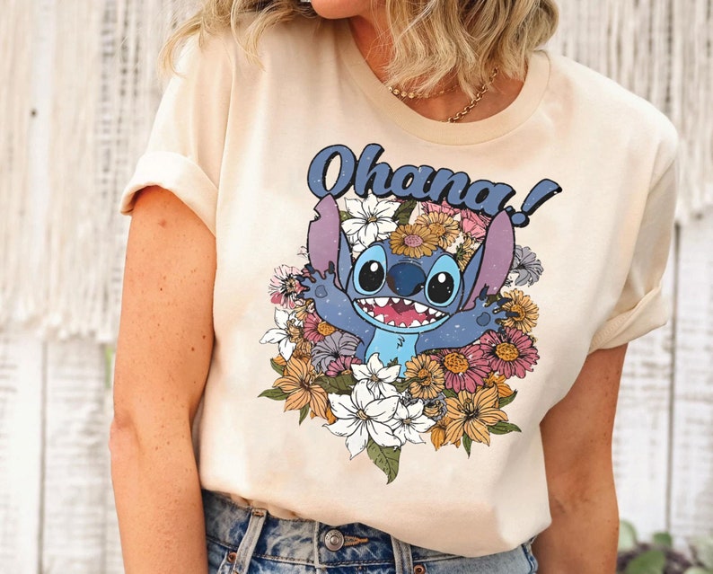 May include: A light beige t-shirt with a blue Stitch character from Lilo & Stitch surrounded by colorful flowers. The text "Ohana!" is printed in black above the character.