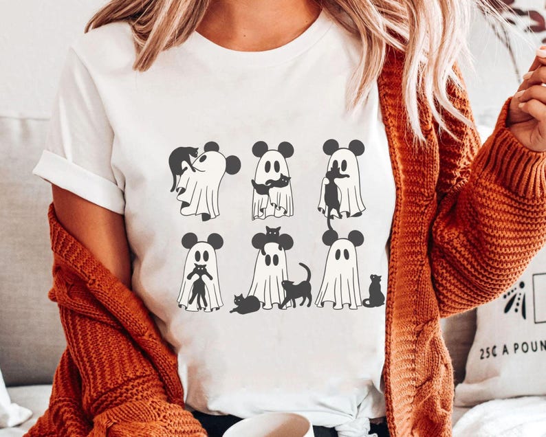 May include: White t-shirt featuring a design of black cats and ghosts wearing Mickey Mouse ears. The ghosts are holding the cats.