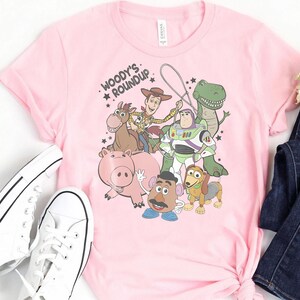 Puede incluir: Camiseta rosa claro con un estampado de personajes de Toy Story. El dise&ntilde;o incluye a Woody, Buzz Lightyear, Rex, Hamm, Sr. Potato, Slinky Dog y Bullseye. El texto "Woody's Roundup" est&aacute; encima de los personajes.