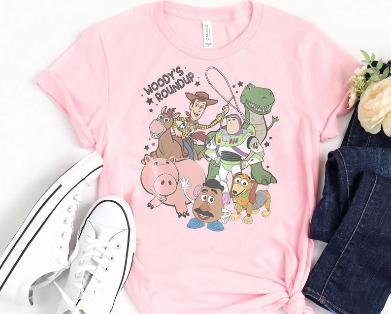 Puede incluir: Camiseta rosa claro con un estampado de personajes de Toy Story. El dise&ntilde;o incluye a Woody, Buzz Lightyear, Rex, Hamm, Sr. Potato, Slinky Dog y Bullseye. El texto "Woody's Roundup" est&aacute; encima de los personajes.