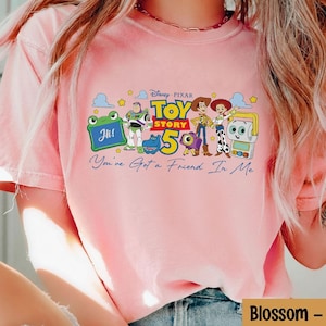 May include: A light pink t-shirt featuring a graphic of characters from the Disney Pixar movie Toy Story. The graphic includes the text "Toy Story 5" and "You've Got a Friend In Me".