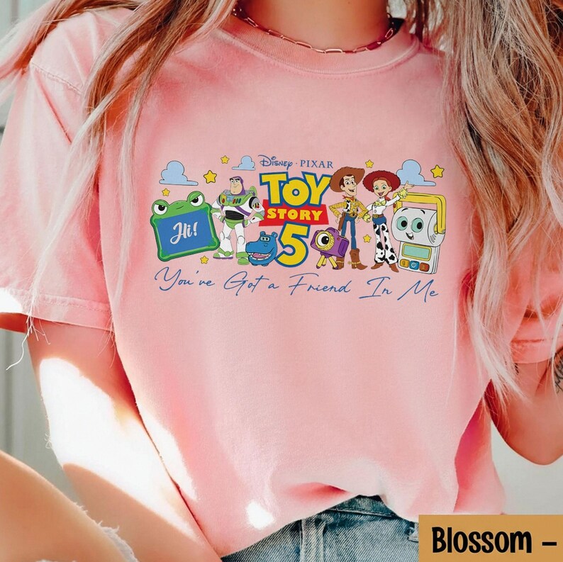 May include: A light pink t-shirt featuring a graphic of characters from the Disney Pixar movie Toy Story. The graphic includes the text "Toy Story 5" and "You've Got a Friend In Me".