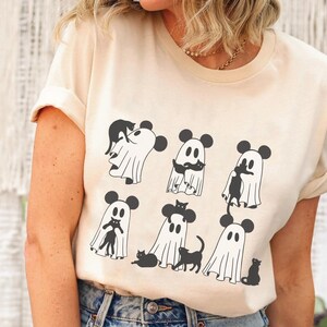 May include: A cream coloured t-shirt featuring a graphic of ghosts wearing Mickey Mouse ears and holding black cats. The ghosts are in various poses, some are holding the cats in their arms, while others are holding the cats by their tails.