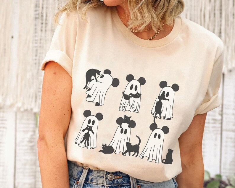 May include: A cream coloured t-shirt featuring a graphic of ghosts wearing Mickey Mouse ears and holding black cats. The ghosts are in various poses, some are holding the cats in their arms, while others are holding the cats by their tails.
