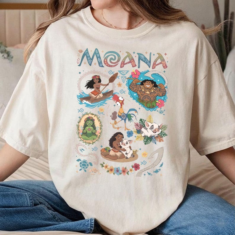 May include: Cream-coloured t-shirt with the word "MOANA" in colourful lettering above cartoon illustrations of characters from the animated film. The illustrations include a woman in a canoe, a muscular man, a green goddess, a rooster, a pig, and a woman with a pig.