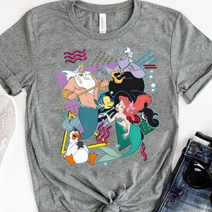 May include: A gray t-shirt with a retro-style graphic of characters from the Disney movie The Little Mermaid. The graphic features Ariel, King Triton, Ursula, and Flounder in a colorful, geometric design.