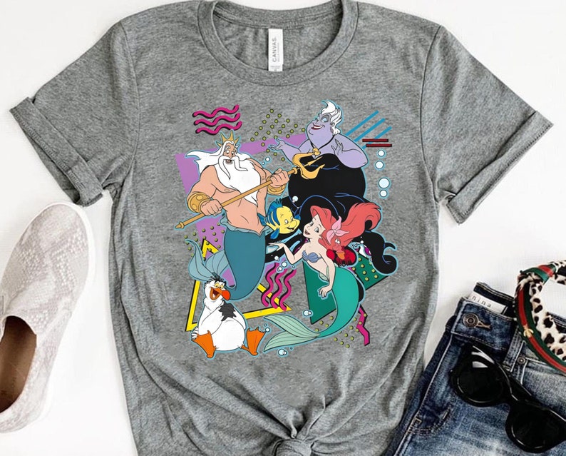 May include: A gray t-shirt with a retro-style graphic of characters from the Disney movie The Little Mermaid. The graphic features Ariel, King Triton, Ursula, and Flounder in a colorful, geometric design.