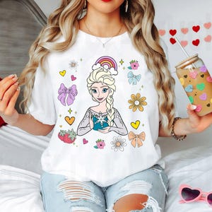 May include: White t-shirt with a colorful cartoon image of a princess with blonde hair and a blue dress. The design also includes a rainbow, flowers, hearts, and bows.