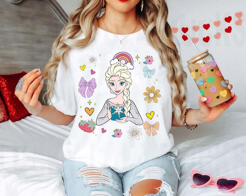 May include: White t-shirt with a colorful cartoon image of a princess with blonde hair and a blue dress. The design also includes a rainbow, flowers, hearts, and bows.