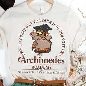 May include: A white t-shirt with a brown owl wearing a graduation cap. The text reads "The best way to learn is by doing it. Archimedes Academy of Wisdom & Wit & Knowledge & Sarcasm."