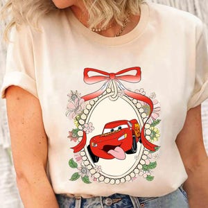Op de afbeelding: Een wit T-shirt met een rode cartoonauto in een decoratief frame. De auto heeft een rood carrosserie, zwarte wielen en een rode strik bovenop. Het frame is versierd met roze en witte bloemen en parels.