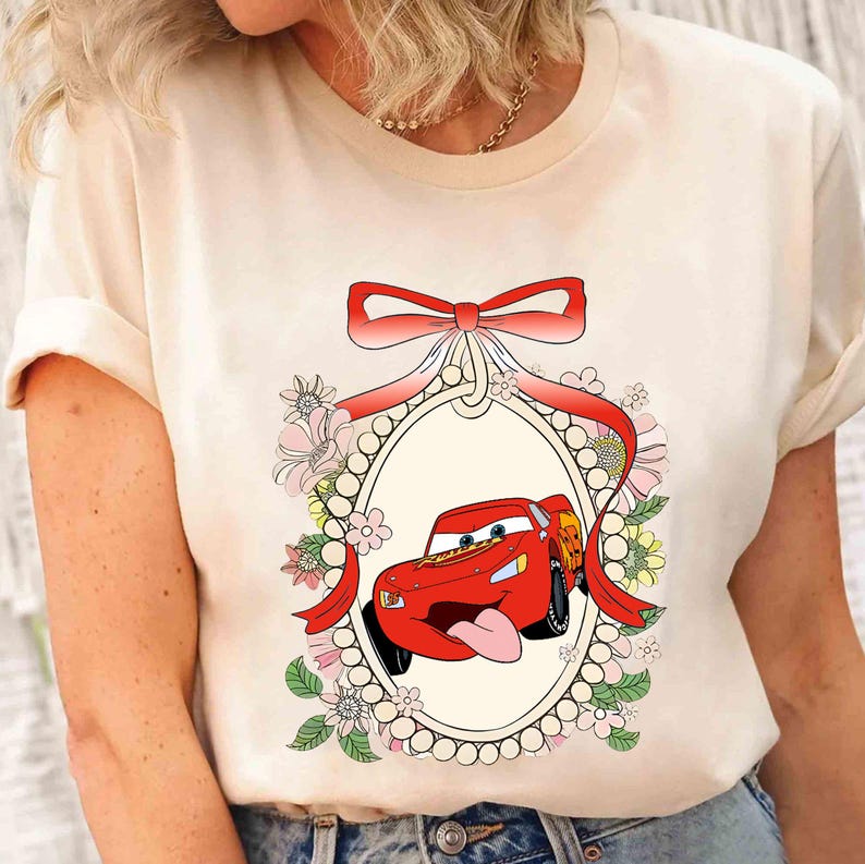 Op de afbeelding: Een wit T-shirt met een rode cartoonauto in een decoratief frame. De auto heeft een rood carrosserie, zwarte wielen en een rode strik bovenop. Het frame is versierd met roze en witte bloemen en parels.