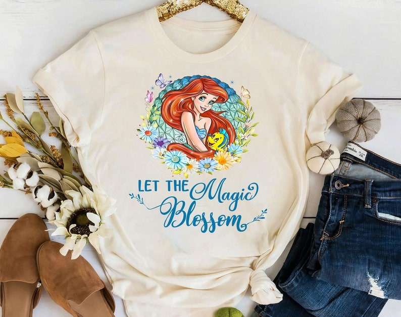 May include: Cream-colored t-shirt featuring a colorful graphic of a red-haired mermaid surrounded by flowers and butterflies. The text "LET THE Magic BLOSSOM" is written in blue script. The shirt is displayed with jeans and fall decor.