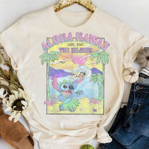 May include: A cream-colored t-shirt with a graphic print of Stitch from Lilo & Stitch. The graphic features Stitch wearing a lei and surfing on a wave. The text "Aloha Hawaii! Come Visit The Islands" is printed above Stitch.