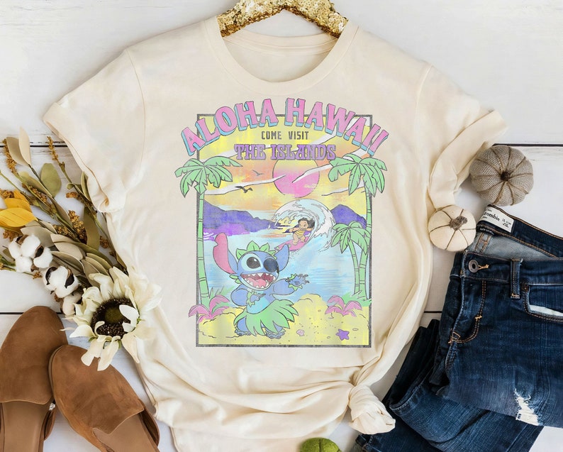 May include: A cream-colored t-shirt with a graphic print of Stitch from Lilo & Stitch. The graphic features Stitch wearing a lei and surfing on a wave. The text "Aloha Hawaii! Come Visit The Islands" is printed above Stitch.