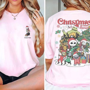 Puede incluir: Camiseta rosa con un gr&aacute;fico de Navidad en la calle principal con personajes de Pesadilla antes de Navidad. El gr&aacute;fico incluye a Jack Skellington, Sally y otros personajes.