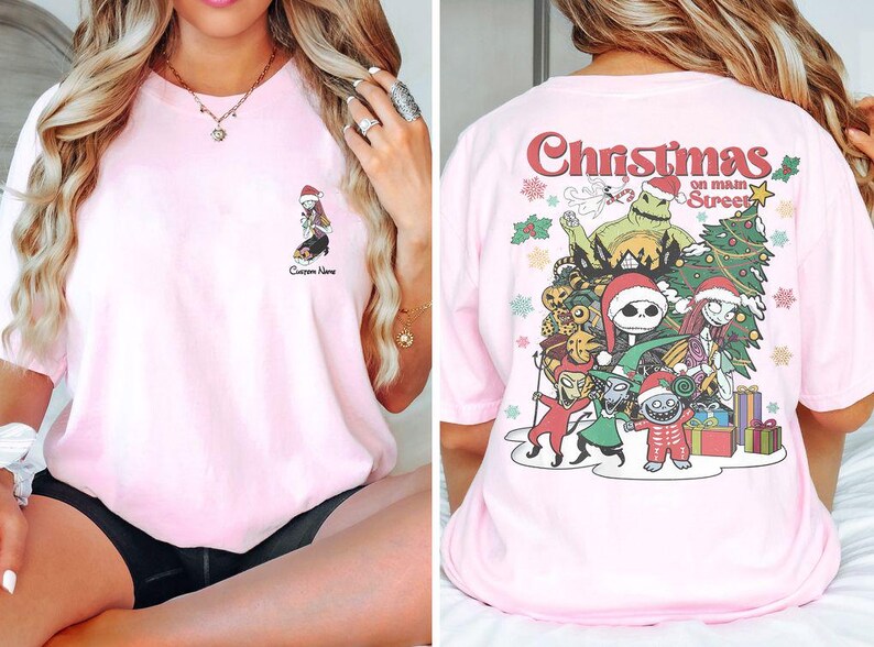 Puede incluir: Camiseta rosa con un gr&aacute;fico de Navidad en la calle principal con personajes de Pesadilla antes de Navidad. El gr&aacute;fico incluye a Jack Skellington, Sally y otros personajes.