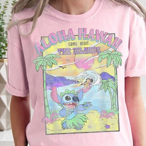 May include: Pink t-shirt with a graphic print of Stitch from Lilo & Stitch surfing in front of a sunset with palm trees and the text "Aloha Hawaii Come Visit The Islands".