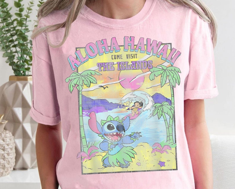 May include: Pink t-shirt with a graphic print of Stitch from Lilo & Stitch surfing in front of a sunset with palm trees and the text "Aloha Hawaii Come Visit The Islands".