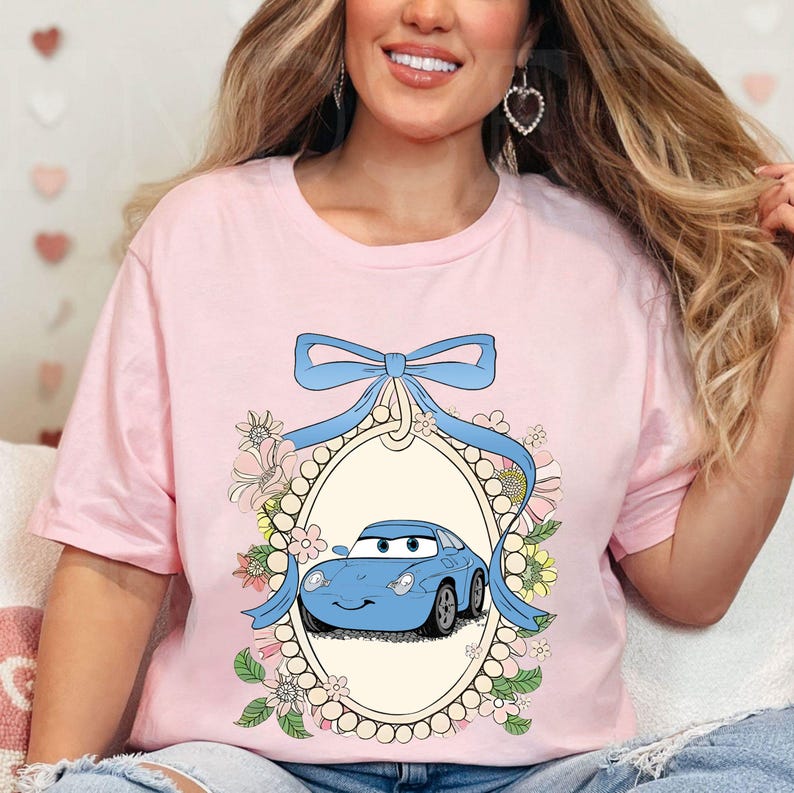 Op de afbeelding: Roze T-shirt met een blauwe auto in een decoratief frame omringd door bloemen en een blauwe strik.