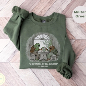 May include: Military green sweatshirt with a vintage illustration of two frogs at a table, with the quote "WE MUST STOP EATING! CRIED TOAD AS HE ATE ANOTHER." The sweatshirt has a crew neck and long sleeves. The design is in a vintage style.