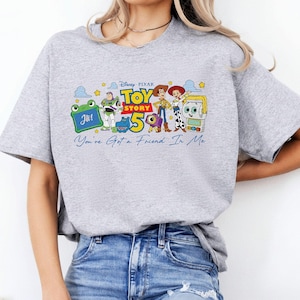 May include: A light gray t-shirt featuring a colorful graphic of characters from the Disney Pixar movie Toy Story. The graphic includes the text "Toy Story 5" and "You've Got a Friend In Me". The shirt has short sleeves and a crew neck.