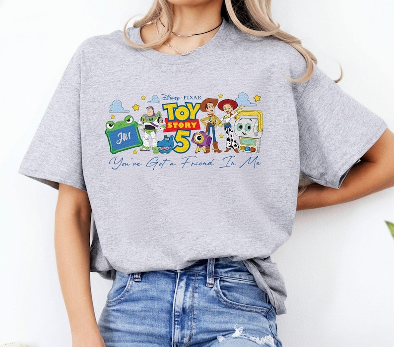 May include: A light gray t-shirt featuring a colorful graphic of characters from the Disney Pixar movie Toy Story. The graphic includes the text "Toy Story 5" and "You've Got a Friend In Me". The shirt has short sleeves and a crew neck.