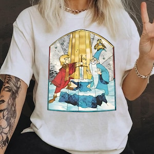 Op de afbeelding: Wit T-shirt met een glas-in-lood ontwerp met een jonge jongen met een zwaard, een tovenaar en een vogel. Het ontwerp is in tinten blauw, geel, rood en groen.