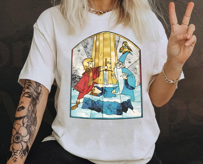 Op de afbeelding: Wit T-shirt met een glas-in-lood ontwerp met een jonge jongen met een zwaard, een tovenaar en een vogel. Het ontwerp is in tinten blauw, geel, rood en groen.