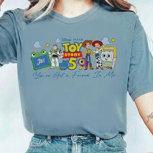 May include: A light blue t-shirt featuring a colorful graphic from the Disney Pixar movie Toy Story 5. The design includes characters like Woody, Buzz Lightyear, and others, with the text "You've Got a Friend in Me."