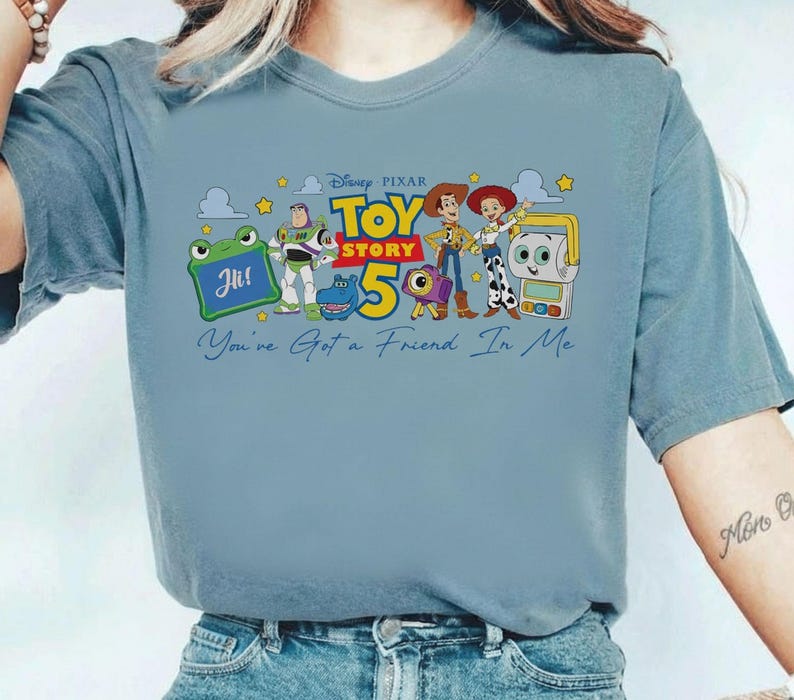 May include: A light blue t-shirt featuring a colorful graphic from the Disney Pixar movie Toy Story 5. The design includes characters like Woody, Buzz Lightyear, and others, with the text "You've Got a Friend in Me."