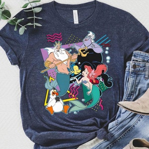 May include: A navy blue t-shirt with a retro-style graphic featuring characters from the Disney movie The Little Mermaid. The graphic includes King Triton, Ariel, Ursula, and Flounder. The graphic is surrounded by geometric shapes and bubbles.