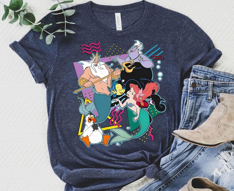 May include: A navy blue t-shirt with a retro-style graphic featuring characters from the Disney movie The Little Mermaid. The graphic includes King Triton, Ariel, Ursula, and Flounder. The graphic is surrounded by geometric shapes and bubbles.