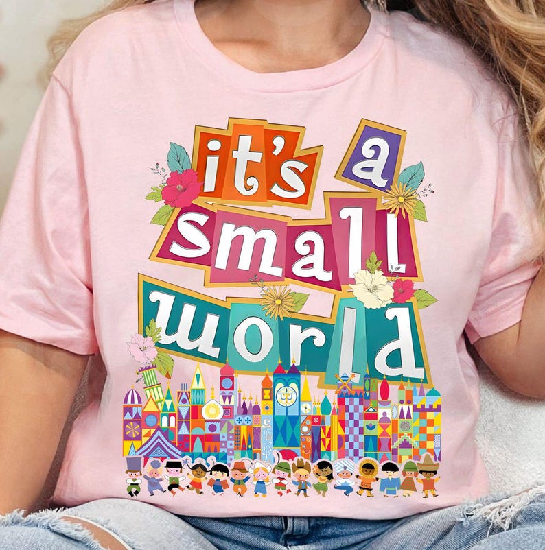 Puede incluir: Camiseta rosa con el texto "it's a small world" en letras de colores. La camiseta presenta un gr&aacute;fico del ic&oacute;nico paseo "it's a small world" en Disney World.