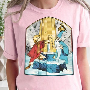 Op de afbeelding: Roze T-shirt met een afbeelding in de stijl van een glas-in-loodraam van een jonge jongen en een tovenaar die een zwaard vasthouden voor een gouden boog. De afbeelding bevat ook een kleine vogel.