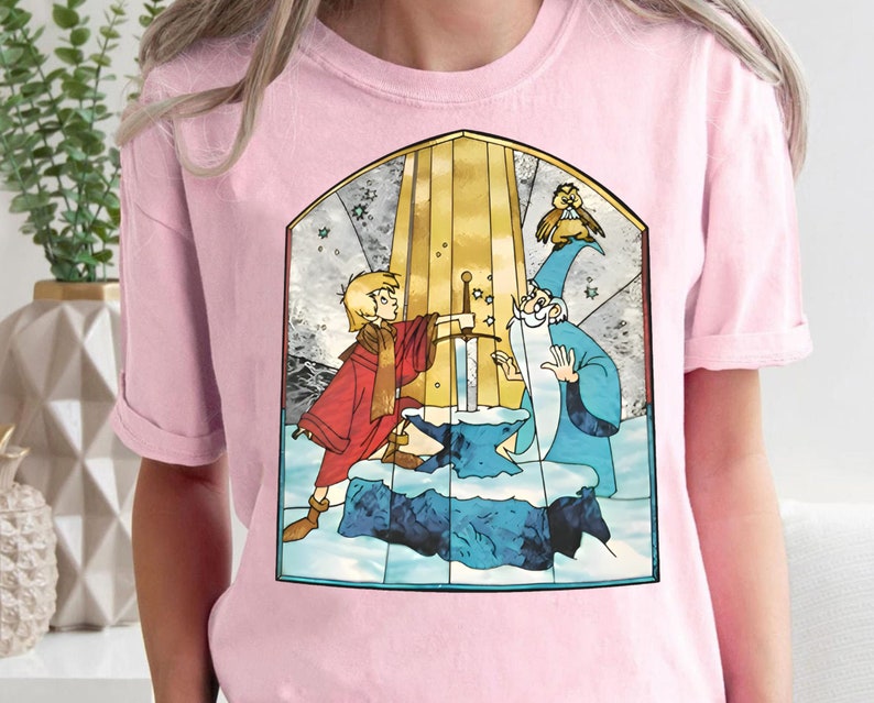 Op de afbeelding: Roze T-shirt met een afbeelding in de stijl van een glas-in-loodraam van een jonge jongen en een tovenaar die een zwaard vasthouden voor een gouden boog. De afbeelding bevat ook een kleine vogel.