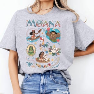May include: A pale grey t-shirt with the word "MOANA" in a decorative font. The shirt displays cartoon illustrations of characters and scenes from the film, including Moana in a canoe and Maui.