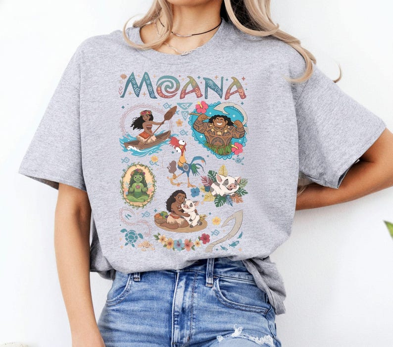 May include: A pale grey t-shirt with the word "MOANA" in a decorative font. The shirt displays cartoon illustrations of characters and scenes from the film, including Moana in a canoe and Maui.