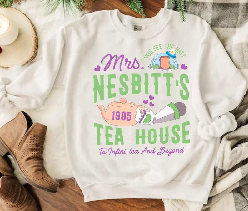K&ouml;nnte beinhalten: Wei&szlig;er Pullover mit einem gr&uuml;nen und violetten Design, auf dem "Mrs. Nesbitt's Tea House" mit einer Zeichentrick-Teekanne und einer Figur aus dem Film Toy Story steht. Der Text lautet auch "You see the hat?" und "1995" und "To Infini-tea And Beyond".
